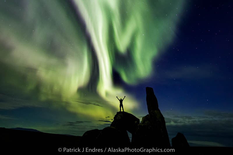 Man stands on a granite tor while the aurora swirls overhead. Bering Land Bridge National Preserve, Seward Peninsula, Alaska.